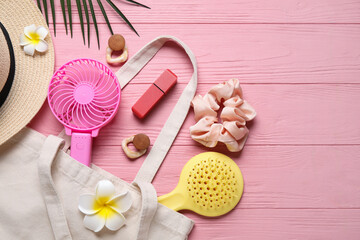 Bag with female summer accessories and mini fan on pink wooden background