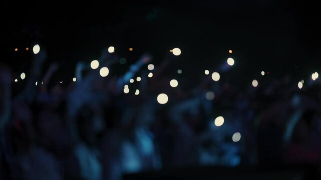 A stunning slow-motion footage of a large crowd at a night concert, waving hands and mobile phones with flashlights in the air. The audience follows the rhythm of the music, creating a magical and emo