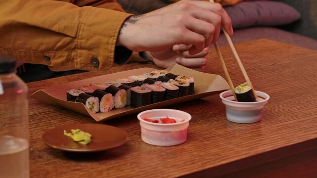Young man sitting at home in evening on the sofa, ordered delivery of tasty asian food, eating sushi rolls with chopsticks, dipping in soy sauce. Slow motion.
