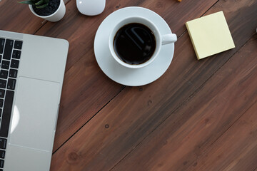 cup of black coffee on white saucer placed on wooden desk alongside laptop, sticky notes, and small potted plant, creating cozy and productive workspace