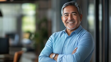 Fototapeta premium Confident middle-aged man in a blue shirt smiling with arms crossed in a modern office setting.