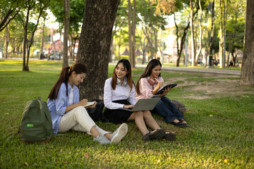Fototapeta premium Three students sitting on the grass, one of them reading a book. The other two are looking at their laptops. Scene is relaxed and casual, as the women are enjoying their time together outdoors 