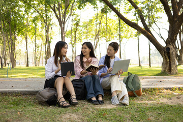 Three students sitting on the grass, one of them reading a book. The other two are looking at their...