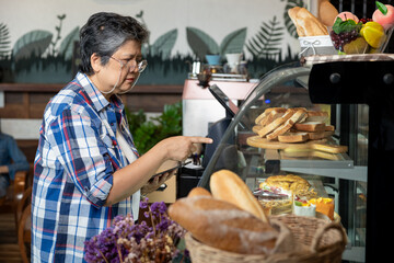 Asian senior woman with short gray hair wearing glasses and plaid shirt standing inside cozy bakery cafe pointing at pastry display case while holding tablet showing active confident in retirement.