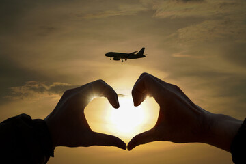 Silhouette of hands forming a heart shape with a passenger plane in the background,sunset background