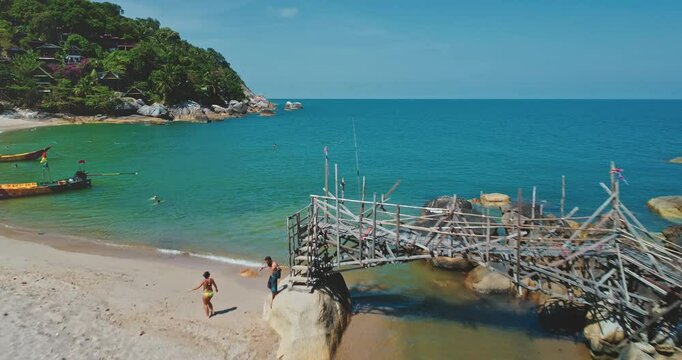 Aerial drone footage capturing a picturesque tropical beach with tourists strolling along the shore, a rickety wooden pier over turquoise waters, and lush greenery on the hillside
