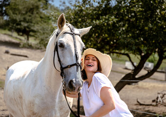 Young caucasian female with white horse in outdoor rural setting, smiling and wearing hat