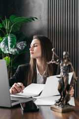 A thoughtful female lawyer works with a laptop and scales of justice on a courtroom table. Concept...