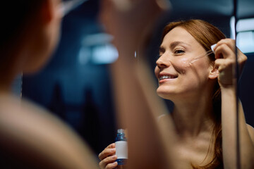 Happy woman using face serum while taking care of her skin in front of mirror.