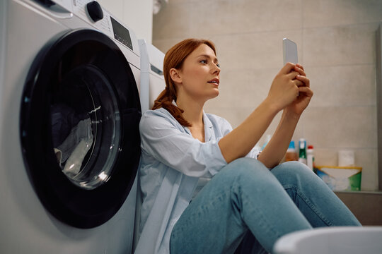 Woman using cell phone while waiting for washing machine to finish in laundry room.