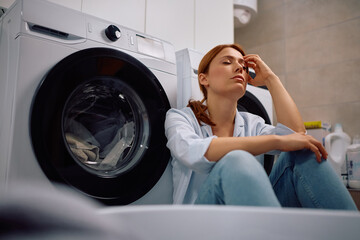 Fototapeta premium Tired woman sitting with eyes closed by washing machine in laundry room.