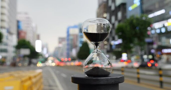 Black sand quickly falling inside hourglass in time-lapse, symbolizing passing time against blurred Gangnam street at background with cars rushing by and city buildings framing busy urban view
