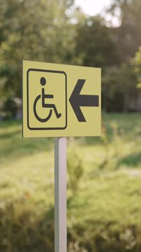 Signage with arrow and wheelchair in park on sunny day. Signage with pictogram for disabled people and their comfort. Infrastructure in public places on vertical video picturesque evening.