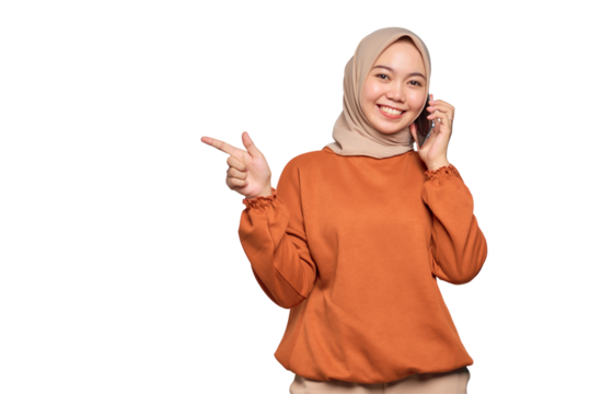 Cheerful young Asian woman in orange shirt talking on mobile phone and pointing finger away at blank space isolated on transparent background