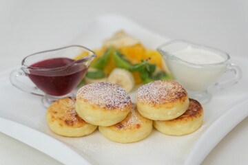Cheese pancakes platter plate featuring three fluffy cheese pancakes, dusted with powdered sugar, accompanied by a dollop of creamy ricotta topped with berry sauce, elegantly arranged on a white dish.