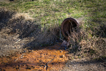 A rusted pipe spills rusty water into a muddy area with weeds and grass growing around it.