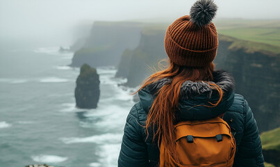 Woman with Red Hair and Backpack Contemplating Dramatic Coastal Cliffs in Misty Weather