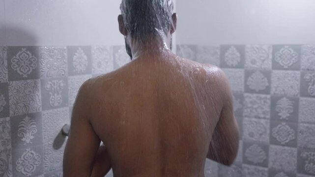 A back shot of a South Asian man rubbing his body with hot water in the shower under bright natural light