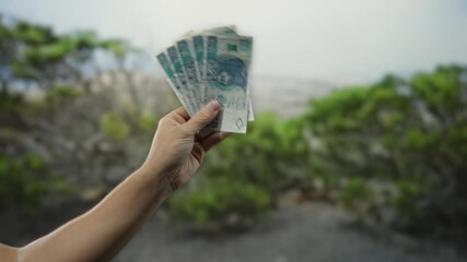 Man holds polish zloty banknotes outdoors in a park with lush greenery, emphasizing finance, currency, and economic themes in a natural setting.