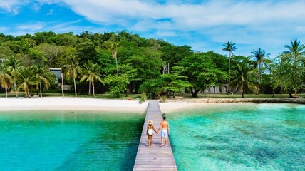 A romantic couple strolls hand in hand along a long wooden pier, surrounded by lush greenery and turquoise waters of Koh Kham Island near Koh Mak Thailand
