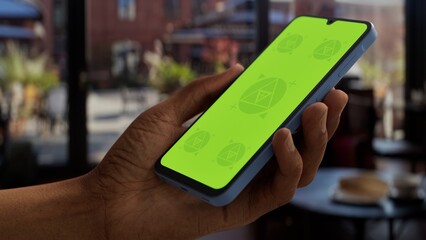 GREEN SCREEN CHROMA KEY A hand holding a smartphone in a bright cafe setting during the day....