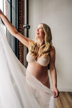 Pregnant woman in lingerie smiles while holding sheer fabric by window