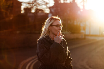 A woman with blonde hair smokes a cigarette at sunset. Golden light shines from behind her.