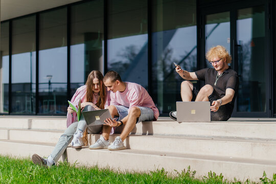 Group of students studying and using laptops outside a modern university building on a sunny day