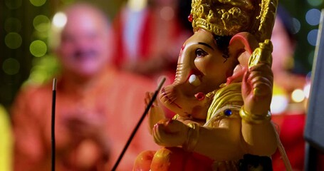 Focus on Lord Ganesha idol during Ganesh Chaturthi as Indian joint family with senior parents, son, daughter-in-law offer prayers with laddu plate and puja thali indoors in decorated festive home