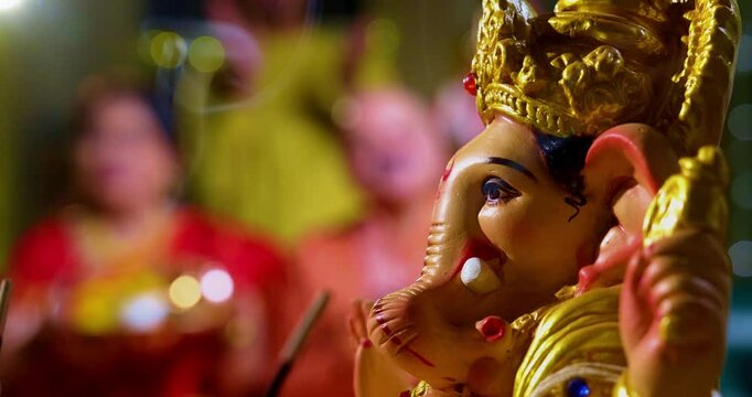 Focus on Lord Ganesha idol during Ganesh Chaturthi as Indian joint family with senior parents, son, daughter-in-law offer prayers with laddu plate and puja thali indoors in decorated festive home