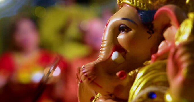 Focus on Lord Ganesha idol during Ganesh Chaturthi as Indian joint family with senior parents, son, daughter-in-law offer prayers with laddu plate and puja thali indoors in decorated festive home