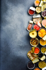 A large assortment of canned preserves food in tin open cans. On a gray stone background. Food banner. Top view.