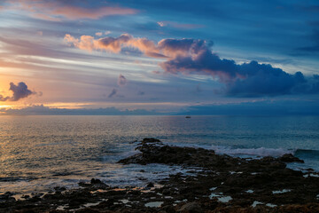 Sunset over rocky coastline with gentle waves and distant sailboat in the serene ocean horizon