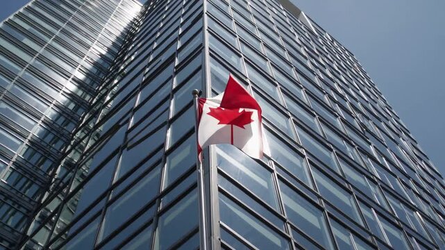 Canadian flag waving on skyscraper against blue sky in urban cityscape.