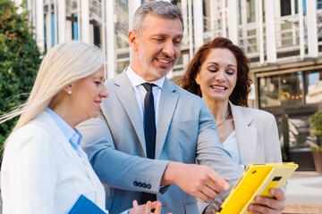 Professional team collaborating outdoors, diverse businesspeople reviewing tablet near contemporary...