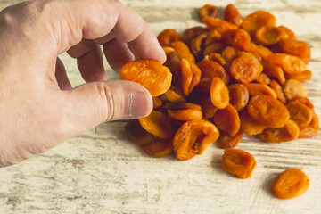  A hand holds one dried apricot against a kitchen table background with a pile of dried apricots,...