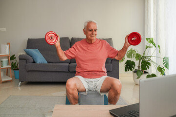 Smiling Senior man exercising with dumbbells at home following online fitness class on laptop