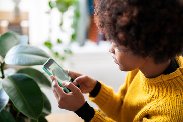 A woman in a yellow sweater uses her phone to photograph a houseplant. She is focused on capturing the perfect shot of the plant's leaves.