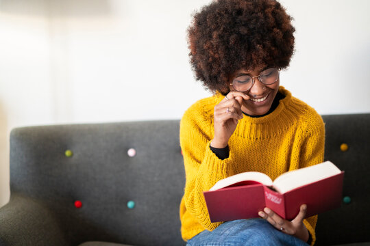 A young woman with glasses and a yellow sweater sits on a couch and reads a book. She is smiling and enjoying the story.