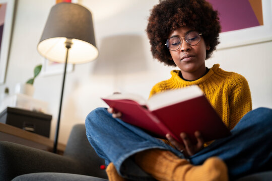 A young woman with glasses and an afro is reading a book. She is sitting comfortably on a couch in a cozy living room, enjoying a quiet moment with her book.