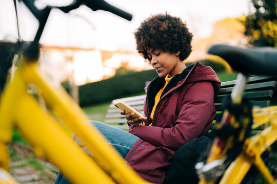 A young woman with an afro is sitting on a park bench, looking at her phone. A yellow bicycle is parked nearby, suggesting she may have been riding.