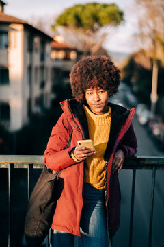 A woman with an afro and a red coat checks her phone on a bridge. She is wearing a yellow sweater and jeans, with a tote bag slung over her shoulder.