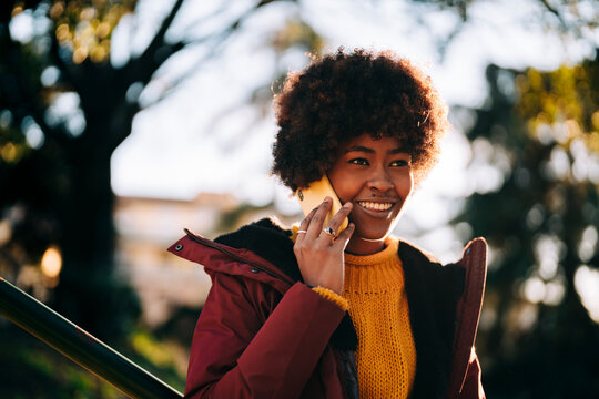 A smiling woman with an afro is outside in a park. She is wearing a winter coat and talking on her cell phone. - Powered by Adobe