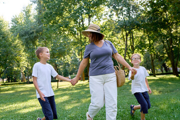 Obraz premium A mom joyfully walks with her two sons in a lush, green park setting under sunny skies, celebrating family bonding and outdoor leisure.
