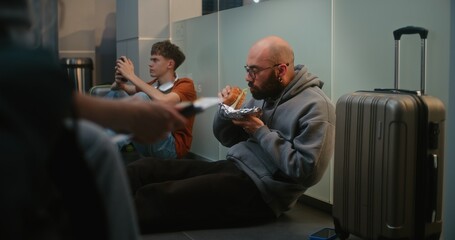 Diverse People and Passengers in Crowded International Airport Terminal: Man Eating Burger, Sitting...
