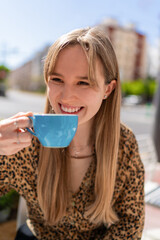 Happy young woman smiling and drinking coffee or tea from a blue cup, sitting at a table in a bar or restaurant in the city during a sunny day
