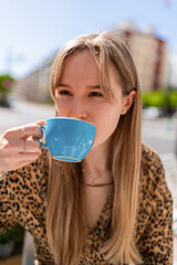 Young woman with long blond hair savoring a cup of coffee from a light blue mug at an outdoor cafe, embracing the vibrant urban atmosphere on a sunny day