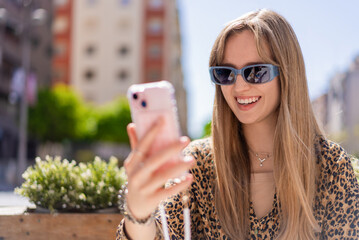 Smiling young woman wearing stylish sunglasses, using a smartphone outdoors in a vibrant urban setting, enjoying sunny weather while connecting with friends through mobile technology