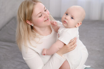 Mom sits on bed and holds baby in arms after waking up, taking care of infant, smiling happily, he feels warm, protected. Concept of motherhood, family, childhood. Mother plays with baby