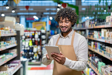 A smiling store clerk checks inventory with a tablet in a grocery store setting. Shelves filled with products surround the employee.
